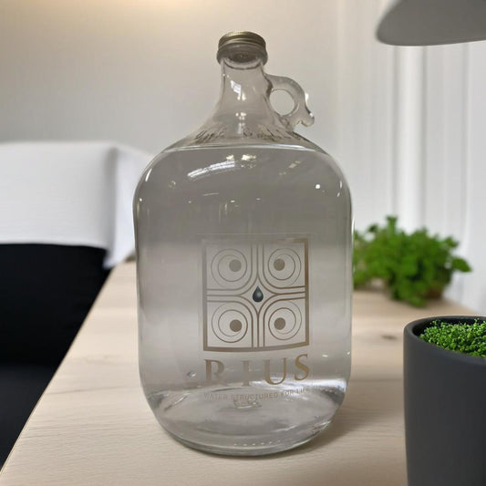 An image of a clear 1 gallon glass bottle of Rius structured water in front of a kitchen sink 